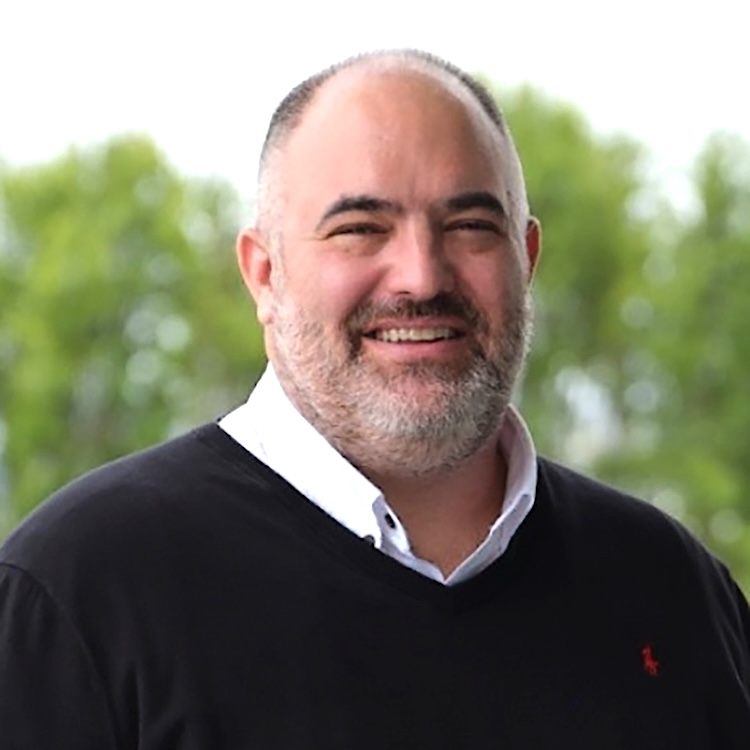 Stephen Gillett. Headshot of man with mustache and beard, wearing dark sweater over a white collared shirt with trees in the background.